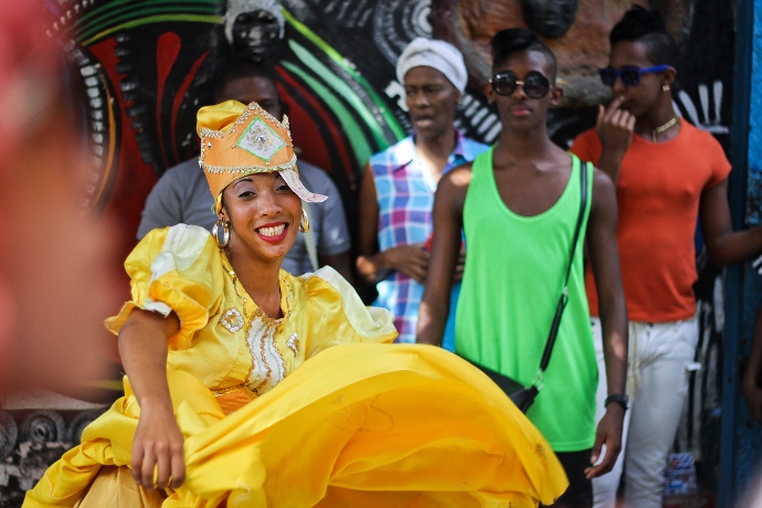 Mulher com roupa carnavalesca amarela posa para foto. Ao fundo outras pessoas a observam dançar