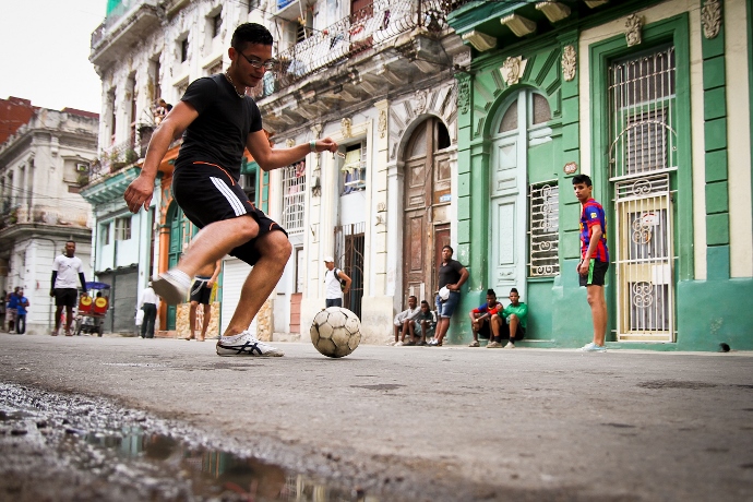 Jovens brincam de futebol na rua. No fundo, outros observam enquanto aguardam encostados na paredes das casas 