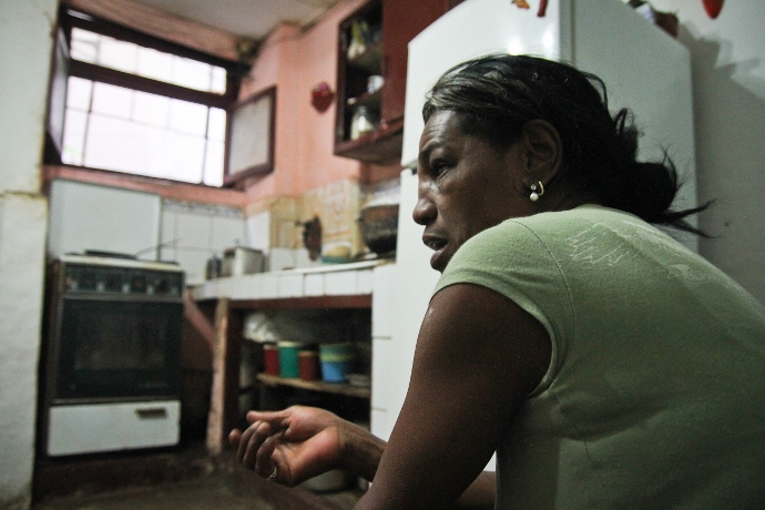 Mulher sentada posa para foto na cozinha de sua casa. Ao fundo, aparecem o fogão e pia com utensílios